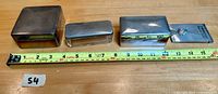 Four vintage metal boxes and razor component lined up with measuring tape for size reference