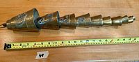 String of nine graduated antique brass cow bells laid out next to a measuring tape showing approximately 20 inches total length. Each bell is embossed with decorative designs and connected on a single rope.