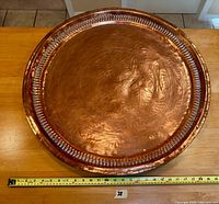 Top view of engraved copper tray showing intricate floral and foliate patterns with a fluted border rim and polished copper surface. Ruler placed below for scale.