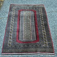 Full view of the Oriental runner rug laid flat on gravel. Shows central red panel with intricate geometric pattern borders in predominantly black, cream, gold, and red hues.