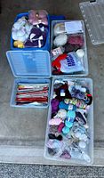 Four clear plastic storage bins containing varied knitting supplies including yarn, needles, and accessories shown from above in an indoor setting.