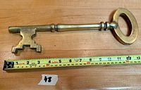 Large antique style brass skeleton key laid flat on wooden surface with tape measure beside it showing length about 12 inches.