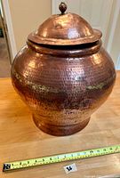 Full view of the large copper storage vessel showing its rounded shape, hammered texture, and lid with finial. Also shows measuring tape for scale (approx. 14 inches wide).