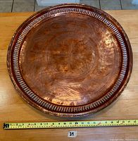 Front view of a large round copper tray with central floral embossing and detailed edge pattern. Measuring tape shows approx. 22 inch diameter.