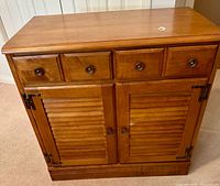Front view of light wood cabinet showing four small drawers with round metal pulls above two closed louvered doors with matching round knobs.
