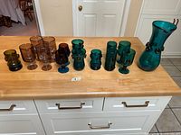 Photo showing entire 13-piece set of mid-century Persian glassware with a large pitcher and assorted glasses in cobalt blue, teal, and amber tones, displayed on a wooden table.