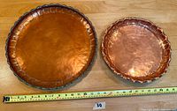 Top view of two round copper trays showing hammered finish and scalloped edges, measuring approximately 18 and 14 inches in diameter with a measuring tape for scale.