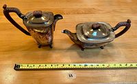 Two vintage silver-plated teapots side by side on wood surface with measuring tape for scale, showing ornate faux wood handles and tarnished surfaces.