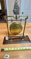 Vintage brass tabletop dinner gong with nautical ship finial and wooden striker. Shown with measuring tape for scale, approximately 10 inches wide base.