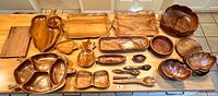 Full overview of more than 30 wooden carved serving pieces displayed on a table including sectioned platters, trays, bowls, and spoons in natural wood finish.