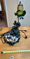 Front view of vintage lamp featuring bronze-tone mermaid figure, shell basin, and green and amber ruffled art glass shade. Electrical cord visible, with measure tape showing approx. 19 inches height.