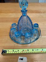 Blue glass decanter with stopper, six matching cordial glasses arranged on clear glass rectangular tray, measuring tape for scale.