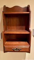 Front view of vintage wooden wall cabinet showing two open shelves and single drawer with brass hardware, wood beadboard back paneling, and scalloped top edge.