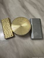Front view of three lighters: gold-tone textured pocket lighter, round gold-tone disc lighter, silver-tone brushed pocket lighter