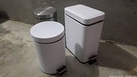 Front view of white round and white rectangular foot pedal bins with stainless bin partially visible