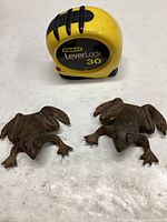 Two cast iron frog figurines shown with tape measure for scale