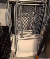 Four folded white plastic chairs stacked, showing grey tubular frames and white seat/back panels