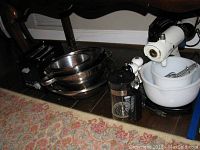 Vintage Sunbeam Mixmaster stand mixer with two white mixing bowls and extra beaters, black 2-slice toaster, stainless steel nested graduated mixing bowls, French press coffee maker, and metal measuring cups grouped on floor near wall.