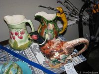Front view of the three pottery items including the pheasant teapot and two pitchers with nature and animal designs on table.