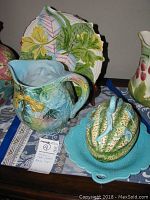 Majolica style pottery pitcher with applied leaves, blue interior, candy dish with lid on scalloped turquoise plate, and leaf-shaped platter behind