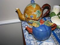Two ceramic teapots on a table with a lace and woven tablecloth, showing detailed colors and design.