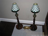 Photo showing two wooden candlesticks with twisted stems, brass accents, and checkered fabric shades alongside a brass pig figurine on the floor.