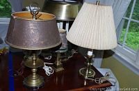 Four assorted vintage lamps on a wooden table including one with a dark decorative metal shade with floral design and black marble base, one with a beige pleated fabric shade, and two with brass and painted glass shades.