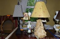 Three assorted lamps on a wooden surface showing a ceramic floral vase lamp, a vintage adjustable arm lamp with checkered shade, and a rooster-based lamp with a chicken wire pattern shade.