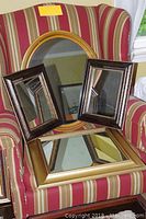 Four mirrors arranged on striped armchair: one large square with gold frame at front, two smaller square dark brown mirrors with beaded edges, one oval mirror with yellow-gold and black striped frame behind.
