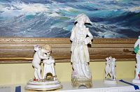 Three porcelain figurines displayed together against backdrop painting. Left cupid sharpening tool, center gentleman, right small cupid grouping.