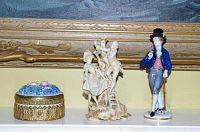 Photo showing two porcelain figurines and a floral trinket box on a white shelf, with visible damage on the seated female figurine.