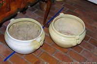 Two round plastic garden planters, light cream color with scroll details, placed on a brick floor near wooden furniture.