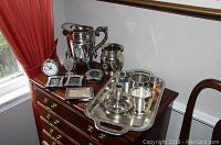 Wide view showing silver plate pitcher, napkin holders, frames, a tray, cup, candle holder, salt shaker, and small clock on wooden chest