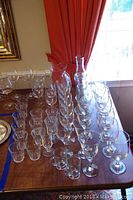 Wide shot of a collection of clear glassware including multiple wine glasses, rock glasses, and two tall pitchers on a wooden table.