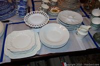 Wide view of assorted ceramic plates, bowls, cups, and creamers on a table showing various designs and colors including white embossed plates and blue patterned dishes.