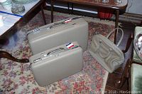 Three American Tourister luggage pieces on a floor next to furniture, including two beige hard-shell suitcases and one soft carry-on bag.