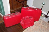 Photo showing three pieces of red American Tourister luggage: two hard shell suitcases and one carry-on bag, positioned on a wooden floor and carpet.