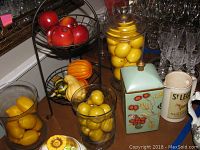 Full view of two-tier metal stand holding faux fruit items including red apples on the top tier and assorted gourds and pears on the bottom tier and surrounding areas with decorative ceramic items nearby.
