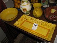 Photo showing large yellow serving platter, yellow bowl, white jar with sunflower painting, and brown fruit-themed bowl.