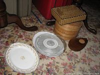 Various ceramic, glass and wood serving platters, bamboo steamer and salt/pepper shaker on carpeted floor near furniture legs.