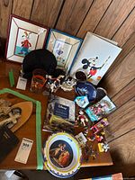Overview of entire lot on tabletop showing framed prints, book, hat, glass, plate, music box, figurines, ornaments and accessories