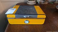 Front view of yellow humidor closed showing carbon-fiber lid panel and hygrometer