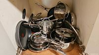 Overview of stacked stainless pots, pans, non-stick frying pans and steamer inserts inside cabinet