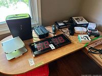 Various electronics on tabletop including LED clock, shredder, laptop stand, phone, surge protector, mini vacuum, stapler, recorder, photo frame box