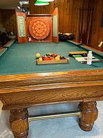 Full view of Olhausen pool table with balls racked, cues visible in corner, dart board cabinet behind