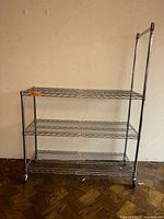 Full view of chrome wire shelving rack showing three shelves, casters, and taller post with hanging bar