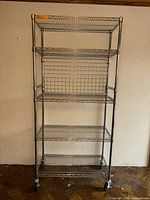 Front view of five-shelf chrome wire rack showing adjustable shelves, grid backing on middle shelf, and caster wheels