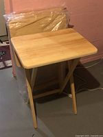 One unpacked wooden folding tray table standing in front of three wrapped tables