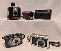 Group view showing five cameras (Kodak Brownie Hawkeye, JVC Everio camcorder, Canon Sure Shot Multi Tele, Sony Cyber-shot, Pentax Optio 750Z) on white surface
