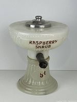 Front view of milk glass dispenser showing Raspberry Shrub lettering, lid and spigot